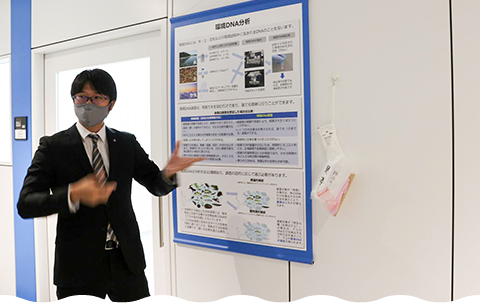 Senior Researcher Shirako giving an explanation in front of an informational poster on environmental DNA analysis